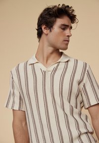 Strikket polo skjorte i kremfarge med vertikale brune striper, med krage og korte ermer, som fremviser teksturert stoff og strukturert design.