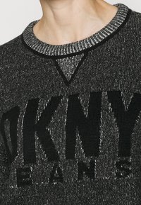 Stickad tröja i mörkgrått med en texturerad yta, med en svart logotyp och en kontrasterande ribbad halsringning.