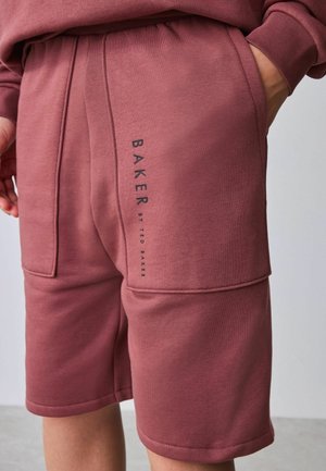 Person trägt mauvefarbene, knielange Shorts und passenden Sweatshirt, Hand in der Tasche, mit dem Schriftzug "BAKER BY TED BAKER" auf dem linken Oberschenkel.
