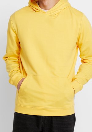 Mies pukeutuneena yksiväriseen keltaiseen huppariin, jossa etutasku ja osittain taskuun laitettuja käsiä, yhdistettyinä mustiin housuihin.