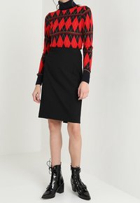 Femme portant un pull argyle rouge et noir, une jupe noire jusqu'au genou et des bottines noires à lacets, se tenant contre un fond blanc uni.