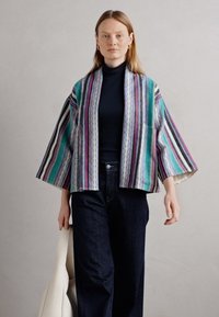 Kimono multicolore à rayures avec de larges manches, fabriqué en tissu doux. Présente des motifs géométriques et une coupe décontractée. Porté par-dessus un haut sombre.