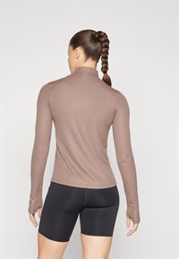 Brown long-sleeve top with a high collar, ribbed texture, and thumbholes, paired with black fitted shorts. Smooth, fitted silhouette.