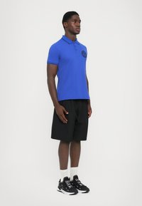 Άνδρας που στέκεται φορώντας μπλε πόλο, μαύρες βερμούδες, λευκές κάλτσες και μαύρα αθλητικά παπούτσια με λευκό κείμενο σε λευκό φόντο.