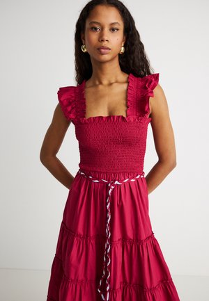 Frau steht mit den Händen hinter dem Rücken, trägt ein ärmelloses rotes Kleid mit gerafften Trägern und geflochtenem Gürtel, goldene Creolen, neutraler Hintergrund.