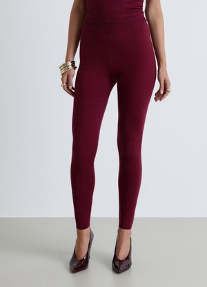 Donna che indossa leggings bordeaux aderenti e top coordinato con décolleté a punta bordeaux scuro e gioielli dorati sulle mani.