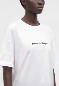 Personne portant un t-shirt blanc avec le texte noir « armani exchange » sur la poitrine, visible des épaules jusqu’au milieu du torse sur un fond uni.