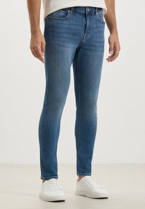 Jeans in denim blu slim con una finitura sfumata, caratterizzati da un classico design a cinque tasche e una vita con passanti per la cintura, abbinati a sneaker bianche.