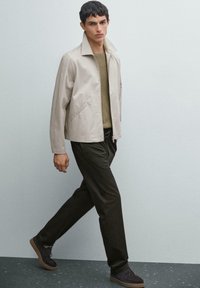 Veste beige clair avec un col haut, chemise kaki en dessous, et pantalon vert foncé. Les chaussures sont sombres avec des semelles brunes.