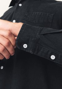 Schwarzes Hemd mit Knopfleiste aus strukturiertem Stoff, mit Brusttasche, weißen Knöpfen und geknöpftem Manschettendetail. Nahaufnahme einer Hand, die die Manschette schließt.