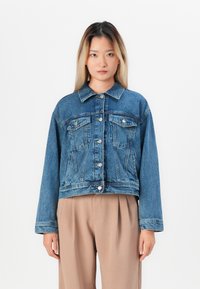 Young woman with straight blonde hair wearing a buttoned blue denim jacket and loose beige pants standing against a white background.