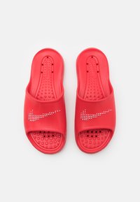 Crvene sandale na klizanje od glatkog plastičnog materijala, s velikim bijelim Nike logotipom od cirkona. Teksturirana unutrašnjost s drenažnim rupama.