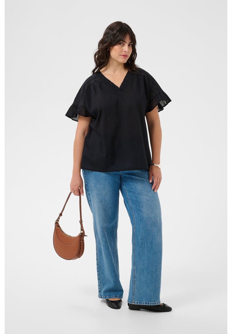 Femme portant un chemisier noir à manches courtes et un jean bleu, tenant un sac à main marron dans sa main gauche sur un fond clair uni.