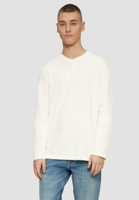 Pikkade varrukatega valge henley särk ümara kaelusega ja viie nupuga, kantud helesiniste teksadega. Pehme, tekstuuriga kangas.
