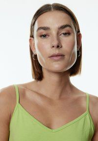Gr�üne Baumwoll-Tanktop mit dünnen Trägern, ausgestattet mit einem V-Ausschnitt und glatter Textur. Das Model zeigt einen neutralen Gesichtsausdruck vor einem weißen Hintergrund.