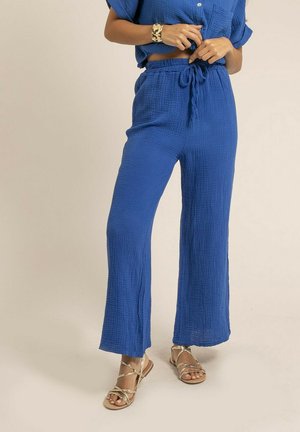 Femme portant un pantalon ample bleu texturé avec une taille à cordon, un haut bleu assorti, un bracelet en or et des sandales dorées à lanières.