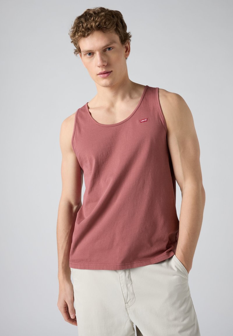Junger Mann trägt ein ärmelloses Tanktop in staubigem Rosa mit kleinem Logo, kombiniert mit hellbeigen Hosen, steht vor einem schlichten Hintergrund.