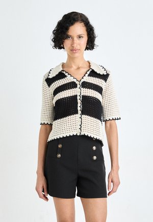 Haut en crochet à manches courtes crème et noir à rayures, avec un col festonné et une fermeture à boutons, associé à un short noir taille haute avec des boutons dorés.