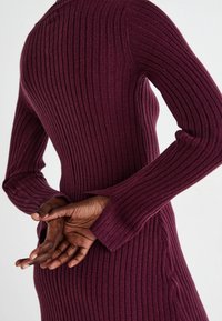 Bordó pletené šaty s dlouhými rukávy, přiléhavým střihem a bočními rozparky. Ruce jsou sepnuté vzadu na spodní části zad, což zvýrazňuje siluetu.