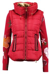 Veste matelassée sans manches rouge avec capuche doublée de fourrure, portée par-dessus de longues manches tricotées rouges à fleurs. Deux poches zippées verticales à l'avant.