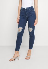 Jeans de mezclilla azul ajustados con desgastes en las rodillas, lavado medio, diseño de talle alto y estilo clásico de cinco bolsillos. Con botón y cremallera plateados.