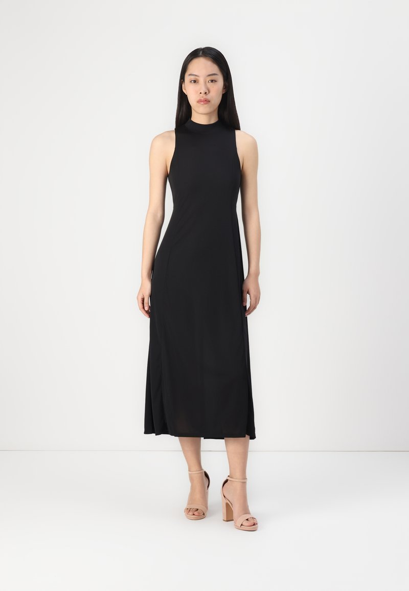 Schwarzes ärmelloses Midikleid aus glattem Stoff, mit einem hohen Kragen und einer fließenden Silhouette. Kombiniert mit beigen, hohen Sandalen.