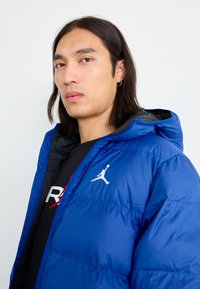 Blå pufferjakke med hette, med et kviltet design og en hvit Jumpman-logo på brystet. Svart innvendig fôr synlig.