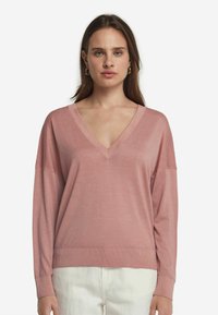 Ljusrosa V-ringad tröja med långa ärmar, mjuk textur och ribbad nederkant. Bärs över vita byxor, vilket framhäver en avslappnad passform.