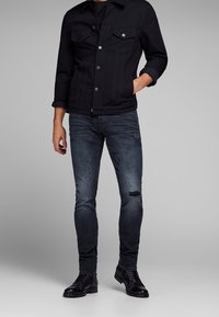 Man med en svart knäppt jacka, mörka slitna skinny jeans och svarta läderstövlar, som står med ena handen i fickan.