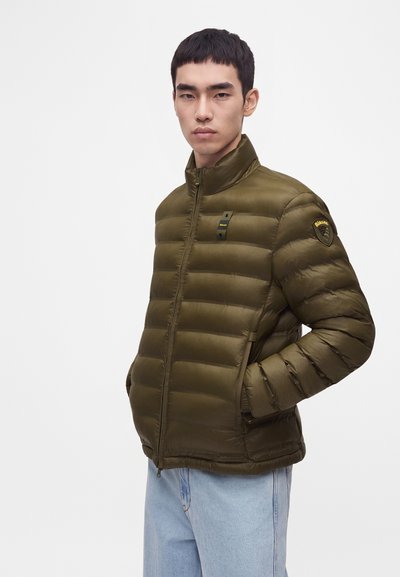 Jeune homme portant une veste matelassée vert olive avec des poches zippées, debout les mains dans les poches devant un fond clair uni.