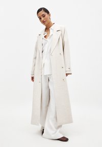Lungo cappotto beige chiaro realizzato in tessuto testurizzato, con una chiusura doppiopetto, colletto oversize e dettagli con bottoni. Abbinato a una camicia bianca e a pantaloni.