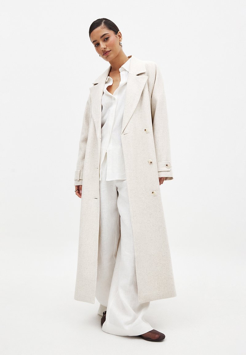 Lungo cappotto beige chiaro realizzato in tessuto testurizzato, con una chiusura doppiopetto, colletto oversize e dettagli con bottoni. Abbinato a una camicia bianca e a pantaloni.