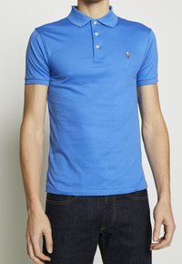 Mies, jolla on päällään istuva sininen polo-paita, jossa on kolme nappia, ja tummat farkut, seisoo yksinkertaista vaaleaa taustaa vasten.