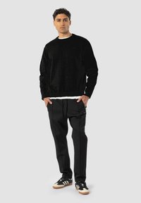 Svart teksturert sweatshirt med rund hals, kombinert med løse svarte bukser og svarte sneakers med beige detaljer. Modellen poserer avslappet.