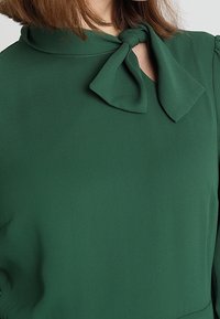 Zelená blůza vyrobená z hladké tkaniny, s vysokým límcem a mašlí. Má dlouhé rukávy a volný střih.