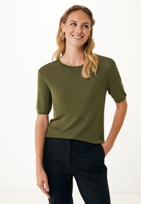 Olivově zelený pletený top s krátkým rukávem, kulatým krajkovým výstřihem a volným střihem. Modelka ho kombinuje s černými kalhotami a rukama v kapsách.