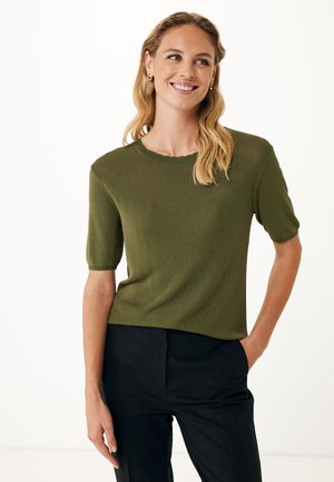 Maglietta a maniche corte in maglia verde oliva con scollatura rotonda a scallop e vestibilità rilassata. Il modello la abbina a pantaloni neri, con le mani in tasca.