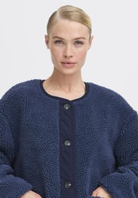 Veste teddy bleu marine en matière polaire texturée, avec patte de boutonnage côtelée et grands boutons noirs, dotée de manches larges.