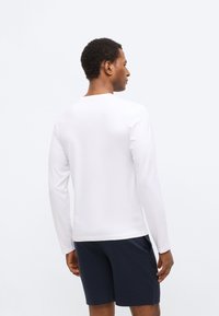 White long-sleeve T-shirt with a round neckline, minimal design, paired with navy shorts. The fabric appears smooth and lightweight.