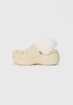 Zuecos beige con un exterior texturizado que cuentan con agujeros de ventilación, un suave cuello de piel sintética blanca y un tacón elevado para mayor comodidad.