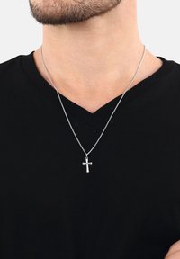 Silberner Kreuzanhänger an einer dünnen Kette, die um den Hals getragen wird, über einem schwarzen V-Ausschnitt-Shirt, das das untere Gesicht und Bartstoppeln zeigt.