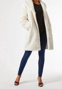 Witte jas van gestructureerd kunstbont met een brede kraag, open voorkant en twee zijzakken, gecombineerd met donkere skinny jeans en zwarte sandalen met bandjes.