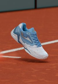 Niebiesko-białe buty sportowe podnoszące czerwoną glinę na korcie tenisowym w pobliżu białej linii.