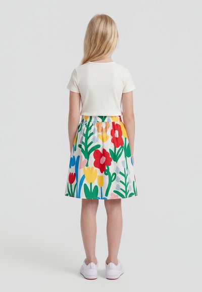 Mladé dievča s blond vlasmi, oblečené v bielom tričku s krátkym rukávom, farebnej kvetinovej sukni a bielych teniskách, stojace čelom odvrátená na jednoliatom pozadí.