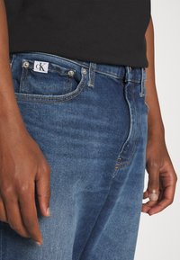 Jeans de mezclilla azul oscuro con una textura sutil, que presentan herrajes plateados, un diseño de cinco bolsillos y una etiqueta con el logo de CK en la cinturilla.