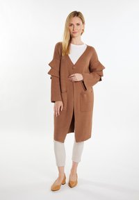 Bruine gebreide cardigan met een V-hals, ruches aan de mouwen, twee voorzakken en een knoopsluiting, gedragen over een wit topje en lichte broek.