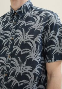 Kurzärmliges Button-Up-Hemd in Marineblau mit einem Muster aus Palmblättern in Hellgrau. Hergestellt aus einem leichten, strukturierten Stoff mit braunen Knöpfen.