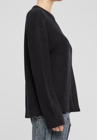 Kobieta w czarnym swetrze z prążkowanej dzianiny z długimi rękawami i okrągłym dekoltem, w zestawieniu z jasnoniebieskimi dżinsami, ukazana z boku.