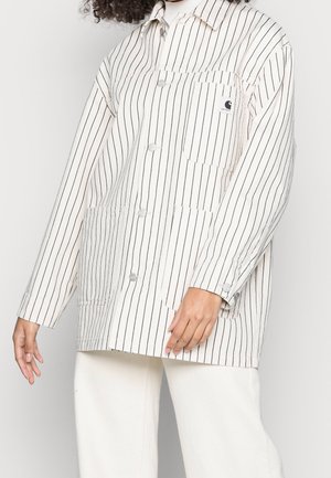 Personne portant une veste blanche boutonnée avec de fines rayures verticales noires et un pantalon assorti, debout devant un fond clair.