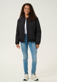 Giacca bomber nera con zip frontale, polsini e orlo a coste. Indossata sopra un top bianco e abbinata a jeans azzurri chiari e sneaker.